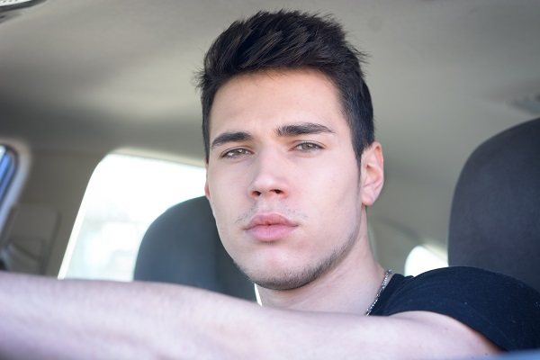 Teenage boy sitting in the driver’s seat of a car, looking forward with a serious expression and his arm resting on the steering wheel.
