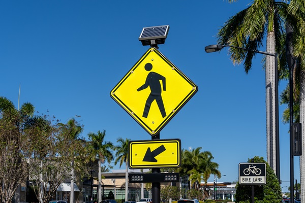 Yellow pedestrian crossing sign with solar panel and arrow, located near palm trees and a bike lane in Florida.