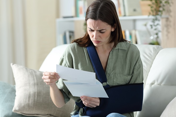 A woman with her arm in a black medical sling sitting on a couch, looking at a letter with a frustrated and confused expression.