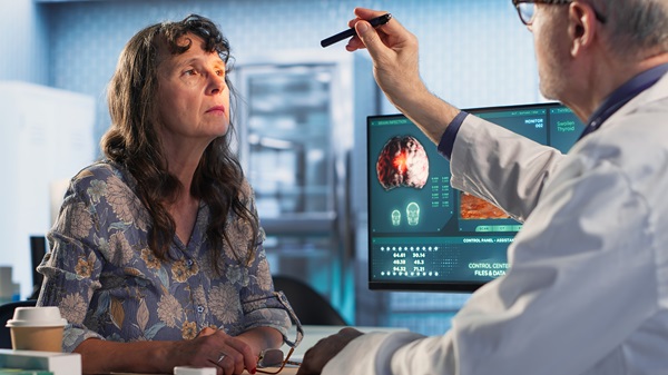 A woman in a floral shirt sits in a medical office in Tavares, FL, as a doctor performs a neurological eye exam with a penlight. A brain scan is visible on a computer monitor in the background.